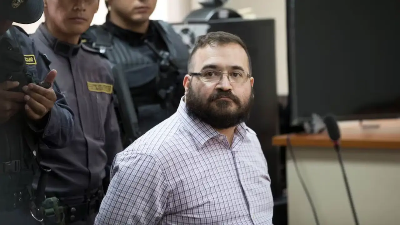 Javier Duarte | México Ahora Bearded man in glasses wearing a light checkered shirt sits in a courtroom with armed officers standing behind him