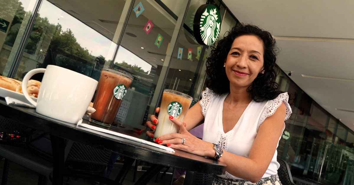 Proyecto nuevo - 2026-04-22T014618535 | México Ahora Woman with curly hair in a white top sits at an outdoor Starbucks table holding a drink with the Starbucks logo