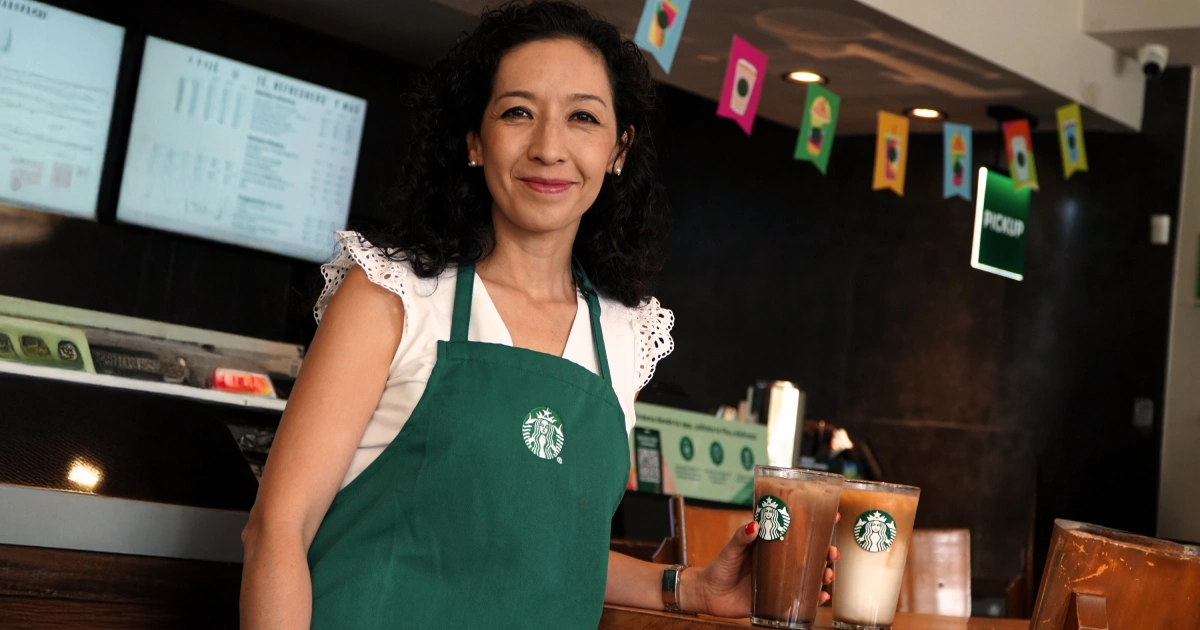 Proyecto nuevo - 2026-04-22T015036252 | México Ahora Smiling barista in a green Starbucks apron holds two drinks at a coffee shop counter