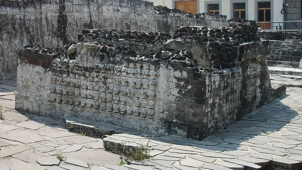 huei-tzompantli-queda-antiguo-estante | México Ahora Weathered stone ruins on a paved plaza a partially collapsed rectangular stone structure with worn blocks and surrounding steps in the background