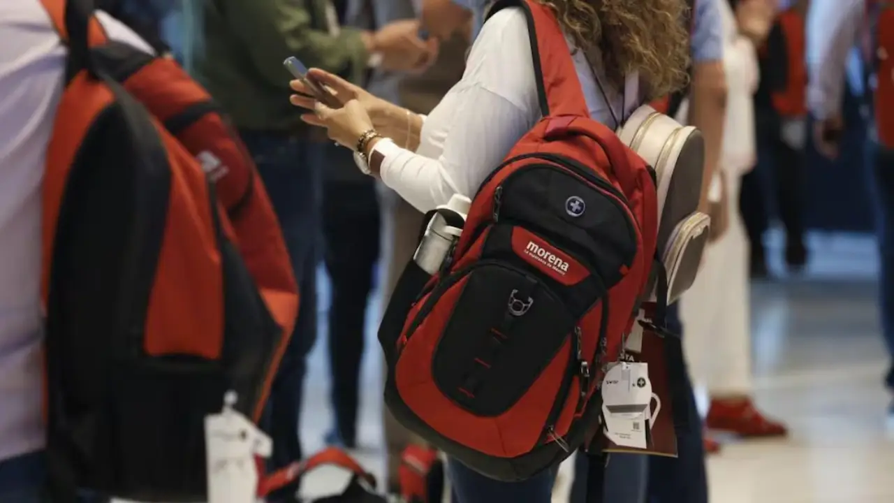 souvenirs de amlo 2 | México Ahora Person wearing a red and black backpack checks a smartphone in a crowded indoor hallway