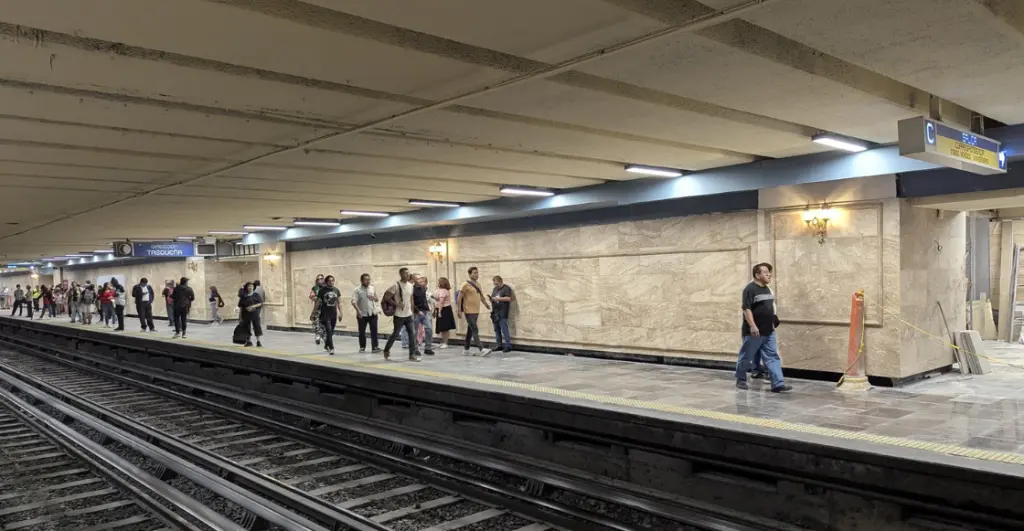 faroles del metro hidalgo | México Ahora Underground subway platform with tracks on the left and a line of people walking and waiting along the edge marble walls and overhead lights