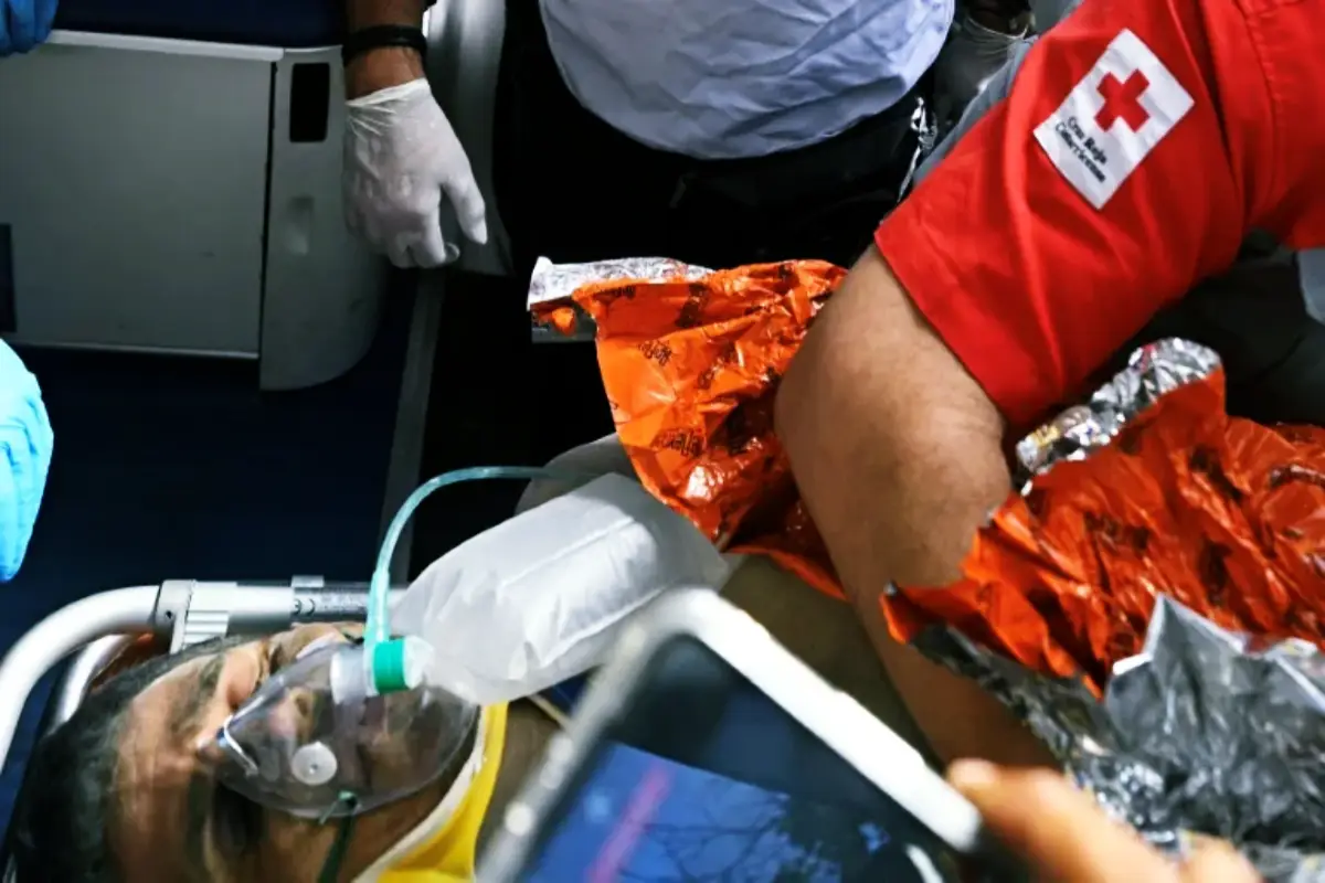 rescatehernangill Tras 100 horas de trabajo rescatan a Hernán en Venezuela con ayuda internacional | Patient on a stretcher wearing an oxygen mask attended by responders with an orange emergency blanket nearby indoors