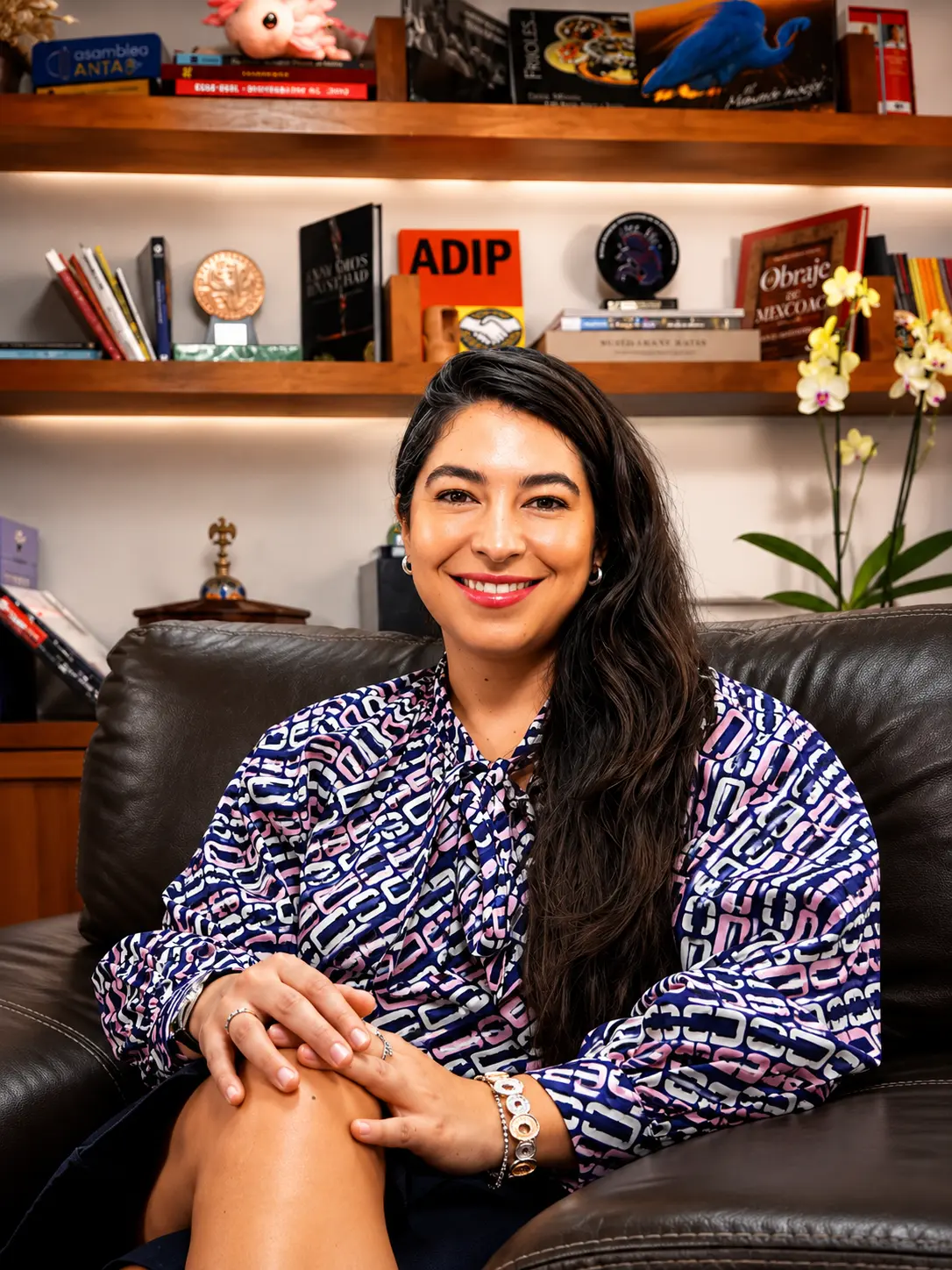 Manola Zabalza Aldama titular de la Secretaría de Desarrollo Económico de la Ciudad de México Mega Clases la apuesta de la CDMX para acelerar la tecnología | Smiling woman with long dark hair sits on a brown leather sofa hands folded with bookshelves in a cozy room behind her