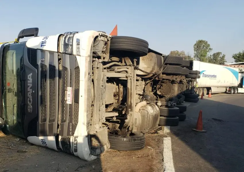 Tráiler vuelca y queda recargado contra barda de piedra