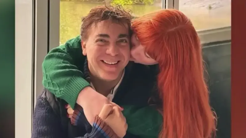 Couple hugging affectionately woman with red hair kisses man on the cheek while he smiles at the camera