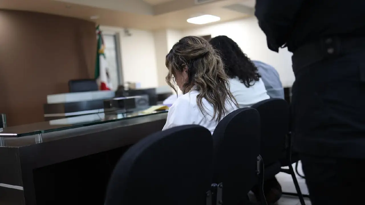 Woman with wavy light brown hair in a white shirt seated at a desk in a formal courtroom or council chamber facing forward