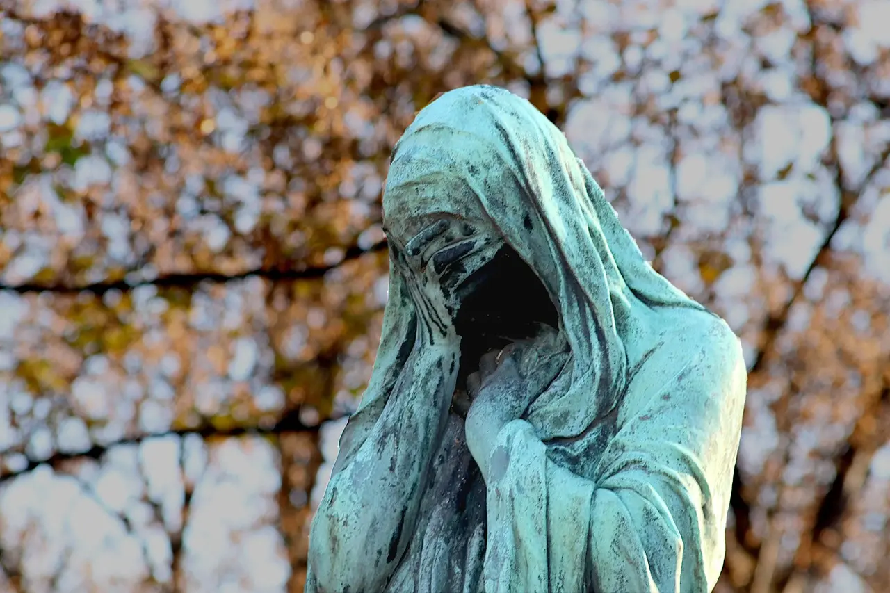 Día Internacional de las Viudas por qué se conmemora este 23 de junio y cuál es su importancia | Bronze statue of a hooded figure covering its face green patina with autumn foliage in the background
