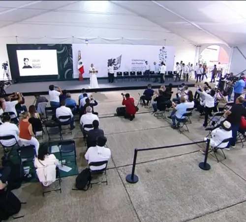 Conference in a white tent with a stage speaker at podium and audience seated facing the stage with cameras filming the event on the sides