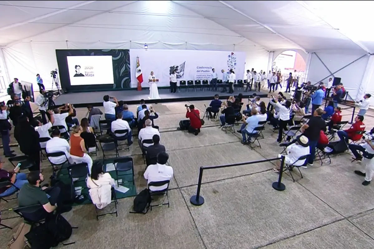 Conference in a white tent with a stage speaker at podium and audience seated facing the stage with cameras filming the event on the sides