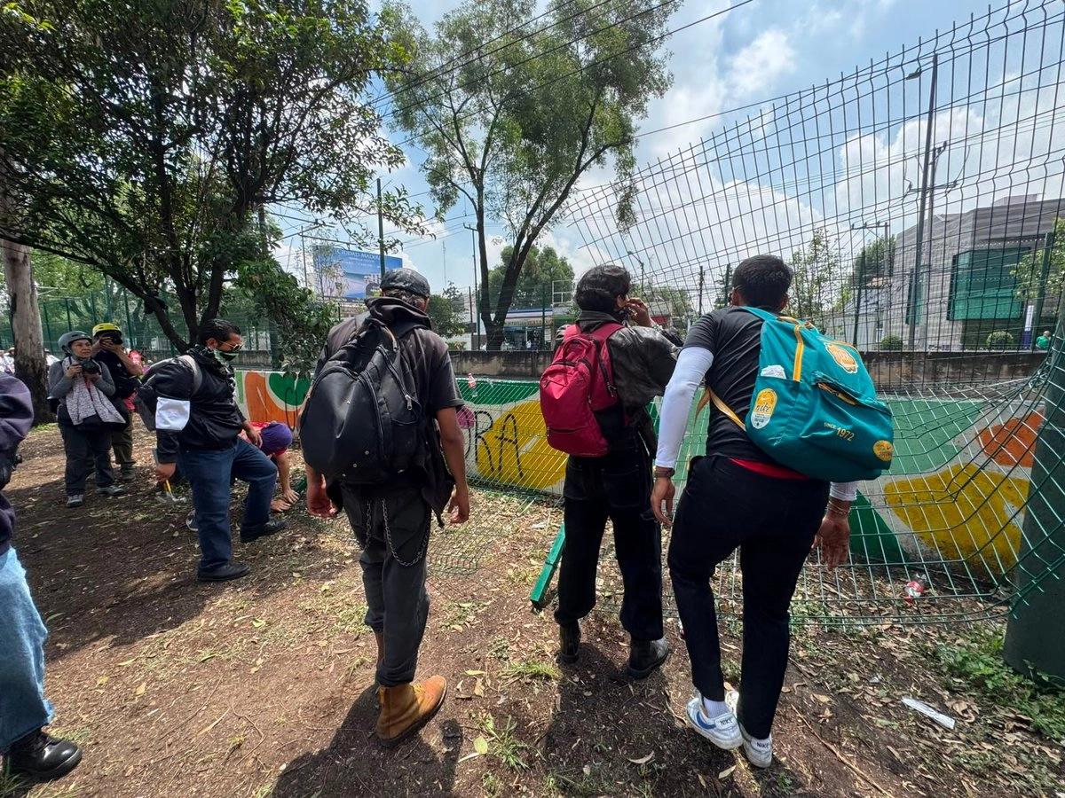 Group of people with backpacks standing along a chain link fence by a small sports court some looking over the fence and one person taking a photo