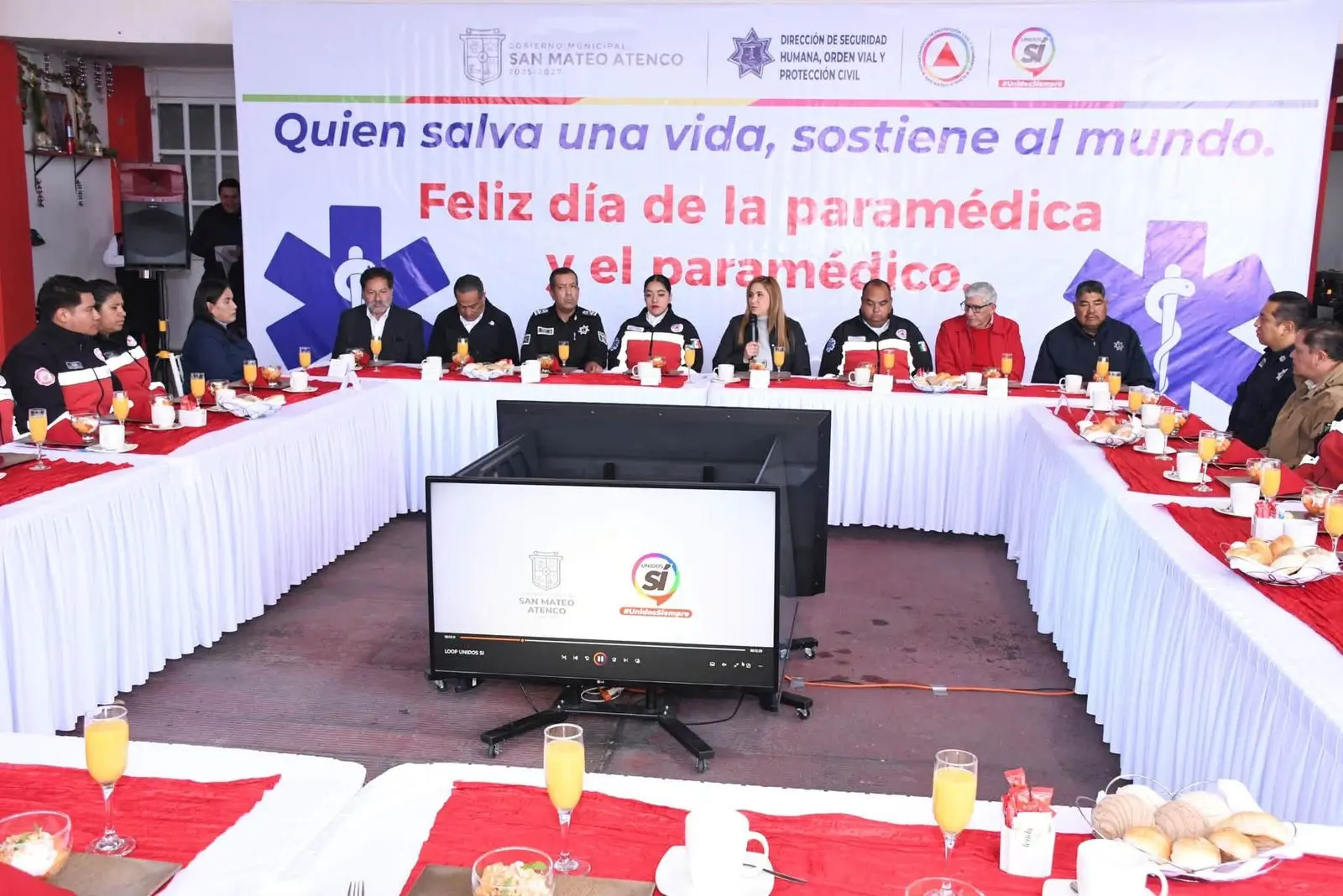 Gobierno de San Mateo Atenco reconoce labor de paramédicas y paramédicos | Grupo de paramédicos y autoridades sentados alrededor de una mesa larga en un evento con un cartel que celebra el día de la paramédica al fondo